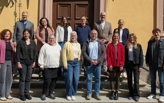Präventionsberaterinnen und -berater Das Bild zeigt eine Gruppe von Personen, die vor einem gelben Gebäude mit einer braunen Tür stehen. Sie sind auf zwei Stufen verteilt und tragen legere bis formelle Kleidung. Die Gruppe scheint für ein Gruppenfoto zu posieren.